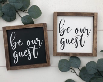 May include: Two wooden signs with the text "be our guest" in black script on a white background and a white script on a black background.