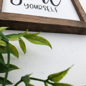 May include: A wooden sign with the text "Be you" and "yourself" in black cursive lettering. The sign is framed in a dark brown wood frame.