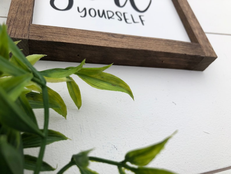 May include: A wooden sign with the text "Be you" and "yourself" in black cursive lettering. The sign is framed in a dark brown wood frame.
