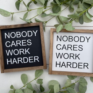 May include: Two wooden signs with the text "Nobody Cares Work Harder" in white lettering on a black background and a white background. The signs are hanging on a white wall.