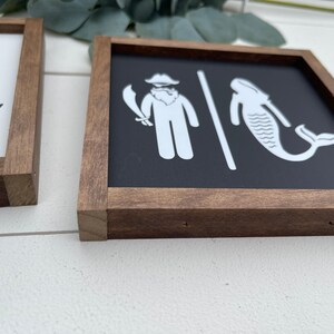 May include: A pair of wooden framed signs with a black background and white illustrations. The sign on the left has a mermaid with a tail and the sign on the right has a pirate with a sword and a whale.