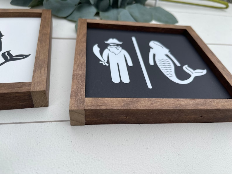 May include: A pair of wooden framed signs with a black background and white illustrations. The sign on the left has a mermaid with a tail and the sign on the right has a pirate with a sword and a whale.