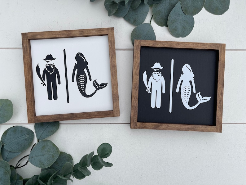 May include: Two wooden signs with a pirate and mermaid design. The signs are black and white and have a rustic wood frame.