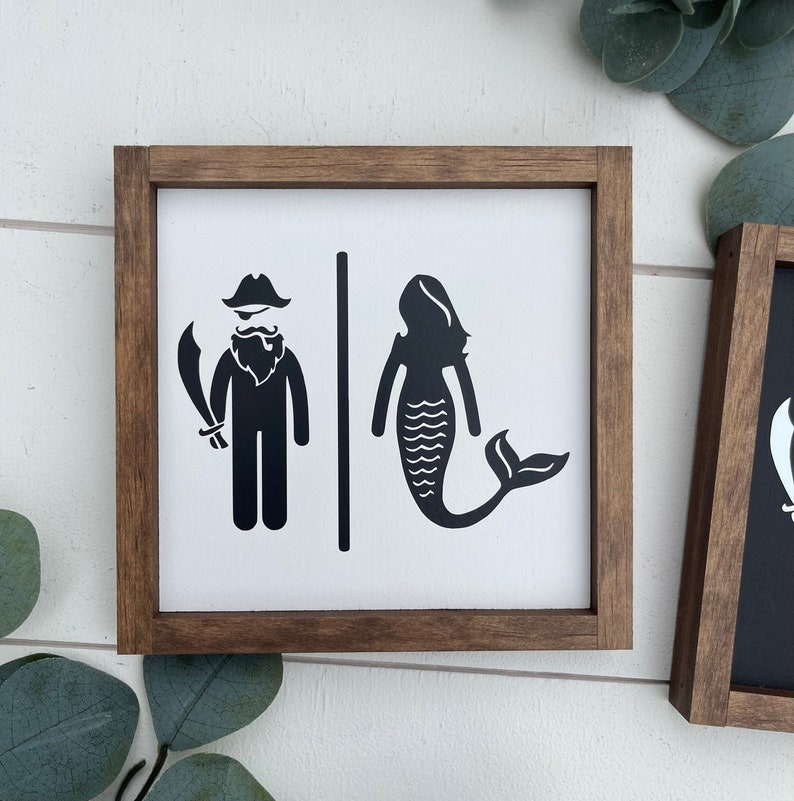 May include: A wooden sign with a white background and black silhouettes of a pirate and a mermaid. The sign reads "Pirate & Mermaid" in black text.