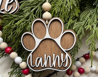 May include: A wooden paw print ornament with the name "Lanny" written in white on it. The ornament is hanging from a string with white wooden beads.
