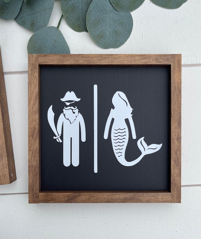 May include: A wooden sign with a black background and white illustrations of a pirate and a mermaid. The sign is framed in a dark brown wood frame.
