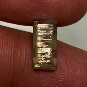 May include: A rectangular, faceted gemstone with a translucent, brownish-grey base colour. The stone features internal, needle-like inclusions that create a distinct, multi-coloured iridescent play of light, displaying flashes of green, pink, and gold.