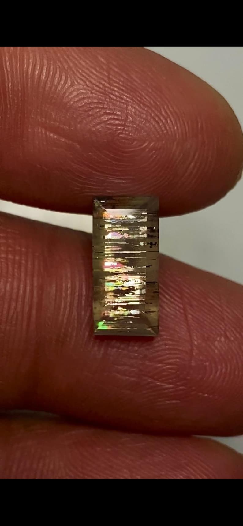 May include: A rectangular, faceted gemstone with a translucent, brownish-grey base colour. The stone features internal, needle-like inclusions that create a distinct, multi-coloured iridescent play of light, displaying flashes of green, pink, and gold.