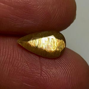 May include: A pear-shaped, faceted gemstone with a metallic, golden-brown lustre and a distinct shimmering, fibrous internal texture. The stone exhibits a reflective surface with multiple cut facets that catch the light, displaying a warm, bronzed hue.