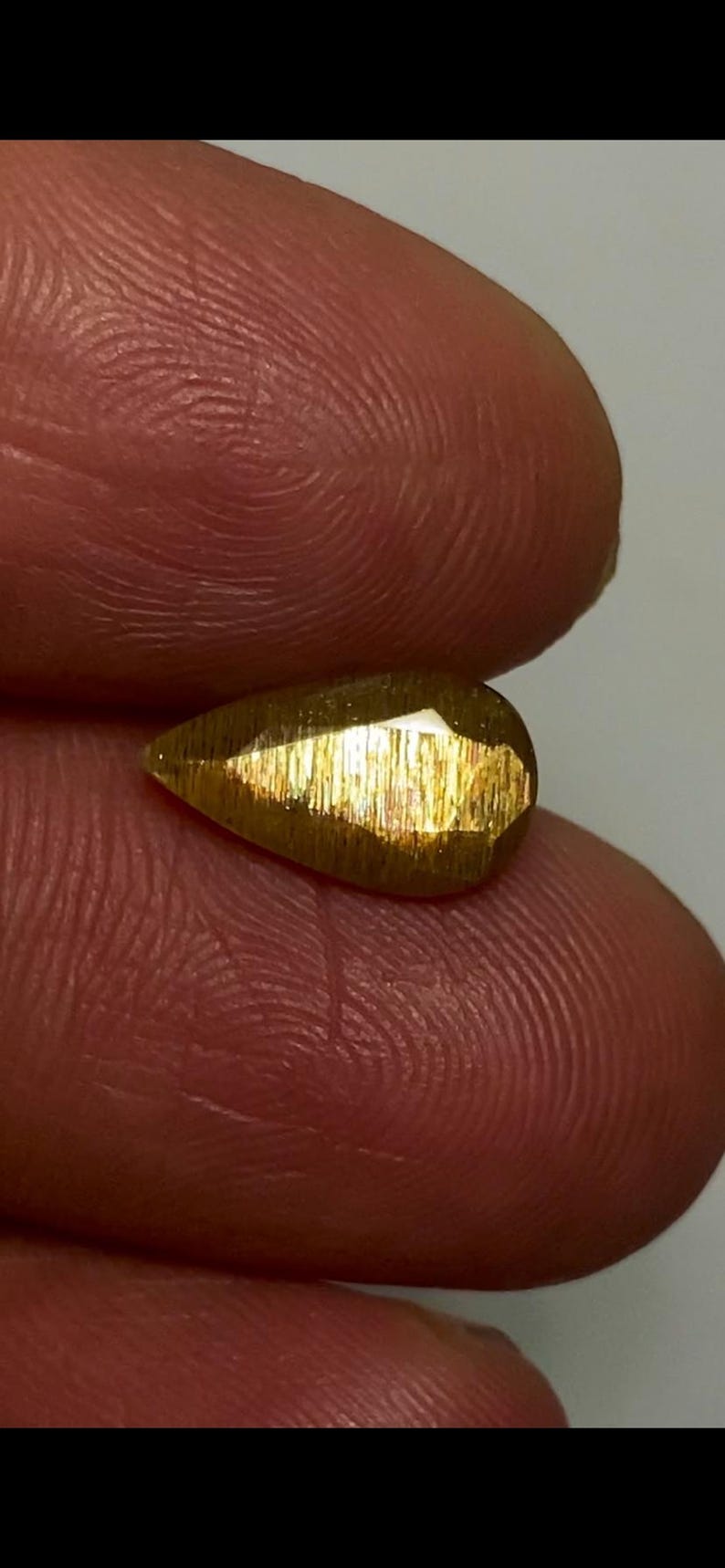 May include: A pear-shaped, faceted gemstone with a metallic, golden-brown lustre and a distinct shimmering, fibrous internal texture. The stone exhibits a reflective surface with multiple cut facets that catch the light, displaying a warm, bronzed hue.