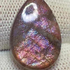 Puede incluir: Una piedra preciosa en forma de lágrima con una superficie brillante e iridiscente. La piedra muestra una mezcla de colores, incluyendo rosa, púrpura y dorado, con un efecto brillante. La gema está sobre un fondo blanco texturizado.