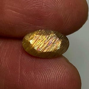 May include: An oval-shaped sunstone gemstone featuring a faceted surface with a golden-brown hue and internal reflective metallic inclusions that create a shimmering effect when light hits the stone's polished, multi-faceted exterior surface.