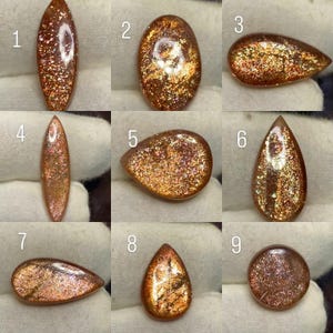 Puede incluir: Nueve cabujones de piedra solar pulidos numerados del 1 al 9, cada uno con un tono naranja-marrón distintivo con inclusiones de brillo metálico. Las formas incluyen cortes marquesa, ovalados, de pera y redondos, todos con superficie brillante.