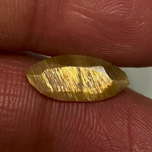 May include: A marquise-cut sunstone gemstone featuring a translucent golden-brown hue with internal metallic copper-coloured inclusions that create a shimmering aventurescence effect across the faceted surface of the stone.