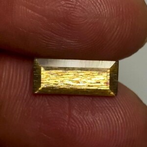 May include: A rectangular gemstone with a faceted edge and a textured, metallic gold-toned surface. The stone features a horizontal orientation with a smooth, reflective top border and a shimmering, granular interior pattern that catches the light.