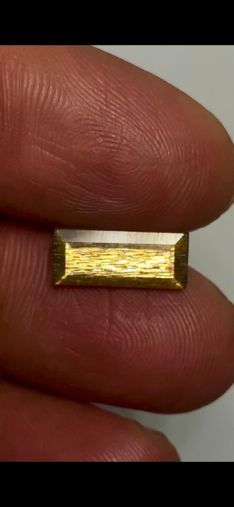 May include: A rectangular gemstone with a faceted edge and a textured, metallic gold-toned surface. The stone features a horizontal orientation with a smooth, reflective top border and a shimmering, granular interior pattern that catches the light.
