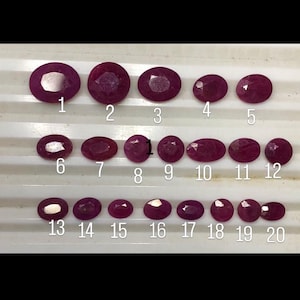 May include: A collection of twenty oval-cut, deep magenta-colored gemstones arranged in three horizontal rows. Each stone is individually numbered from 1 to 20 in white text, showcasing faceted surfaces that reflect light across their polished tops.