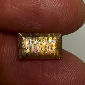 May include: A rectangular sunstone gemstone featuring a faceted surface with internal golden-brown inclusions that display iridescent, multi-coloured flashes of light throughout the stone's transparent, amber-toned crystalline structure.