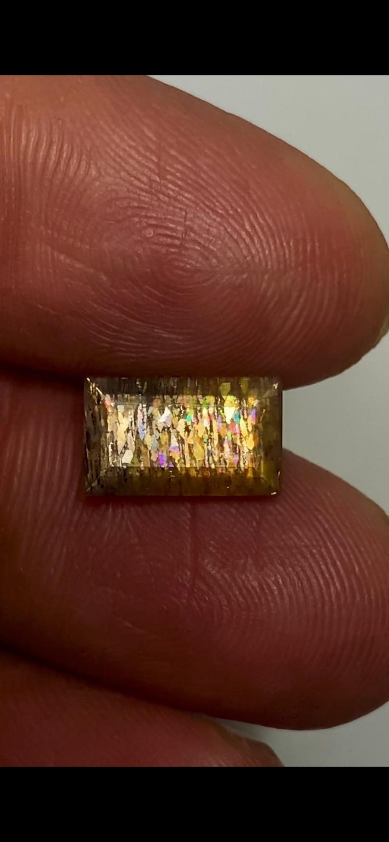 May include: A rectangular sunstone gemstone featuring a faceted surface with internal golden-brown inclusions that display iridescent, multi-coloured flashes of light throughout the stone's transparent, amber-toned crystalline structure.