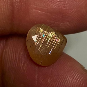 May include: A faceted, translucent golden-brown sunstone gemstone featuring internal metallic inclusions that create a shimmering, reflective effect with subtle flashes of iridescent light across its polished, multi-angled surface.