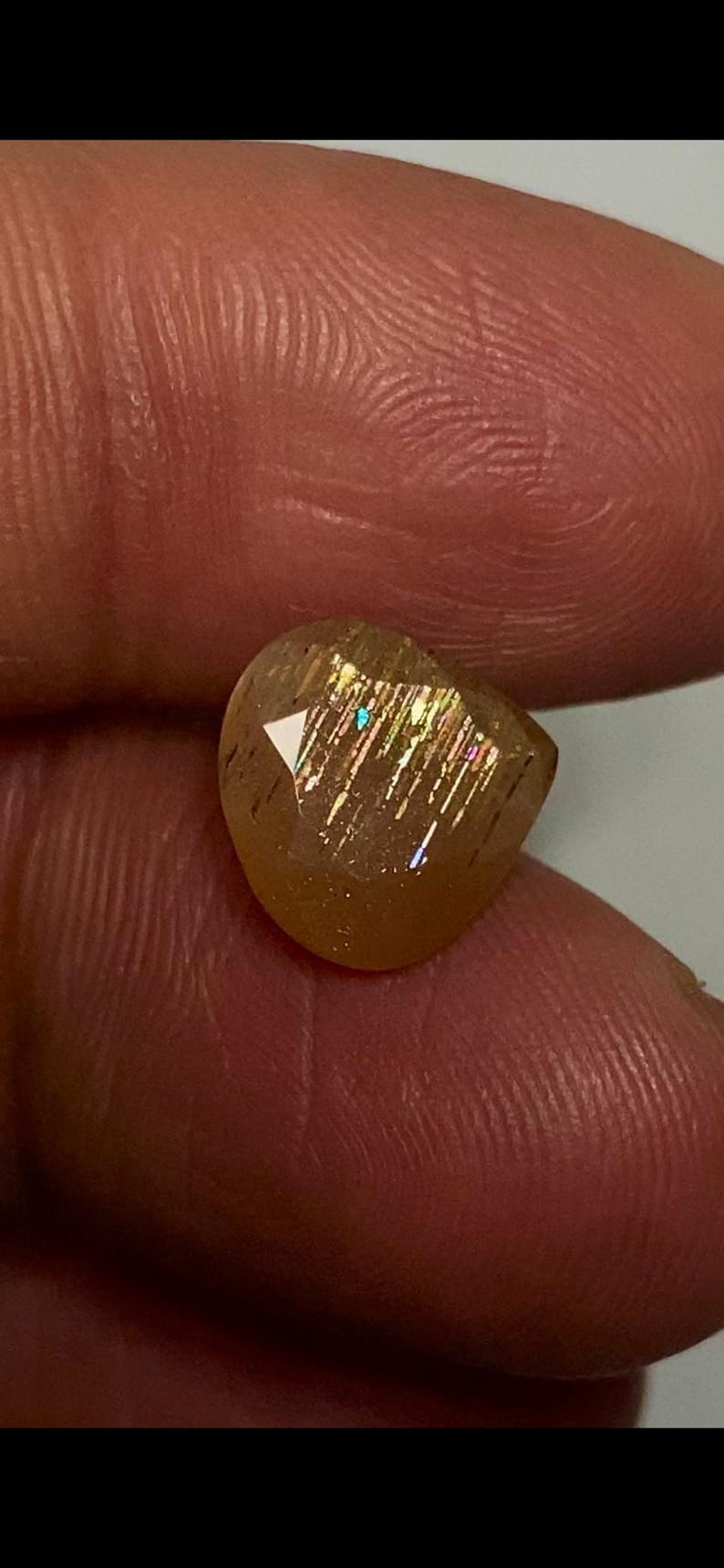 May include: A faceted, translucent golden-brown sunstone gemstone featuring internal metallic inclusions that create a shimmering, reflective effect with subtle flashes of iridescent light across its polished, multi-angled surface.