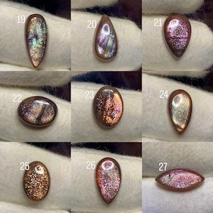 Pode incluir: Uma coleção de nove cabochões de pedra do sol polidos numerados de 19 a 27, cada um apresentando uma base de tom cobre única com inclusões iridescentes e cintilantes que refletem a luz em vários tons de ouro, rosa e azul nas superfícies.