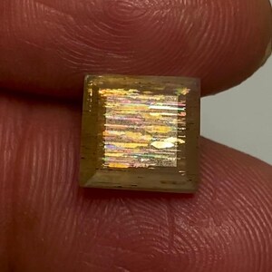 May include: A square-cut sunstone gemstone featuring a translucent brownish-orange body with internal metallic inclusions that create a shimmering, multi-coloured iridescent effect across the centre of the stone's flat, polished surface.