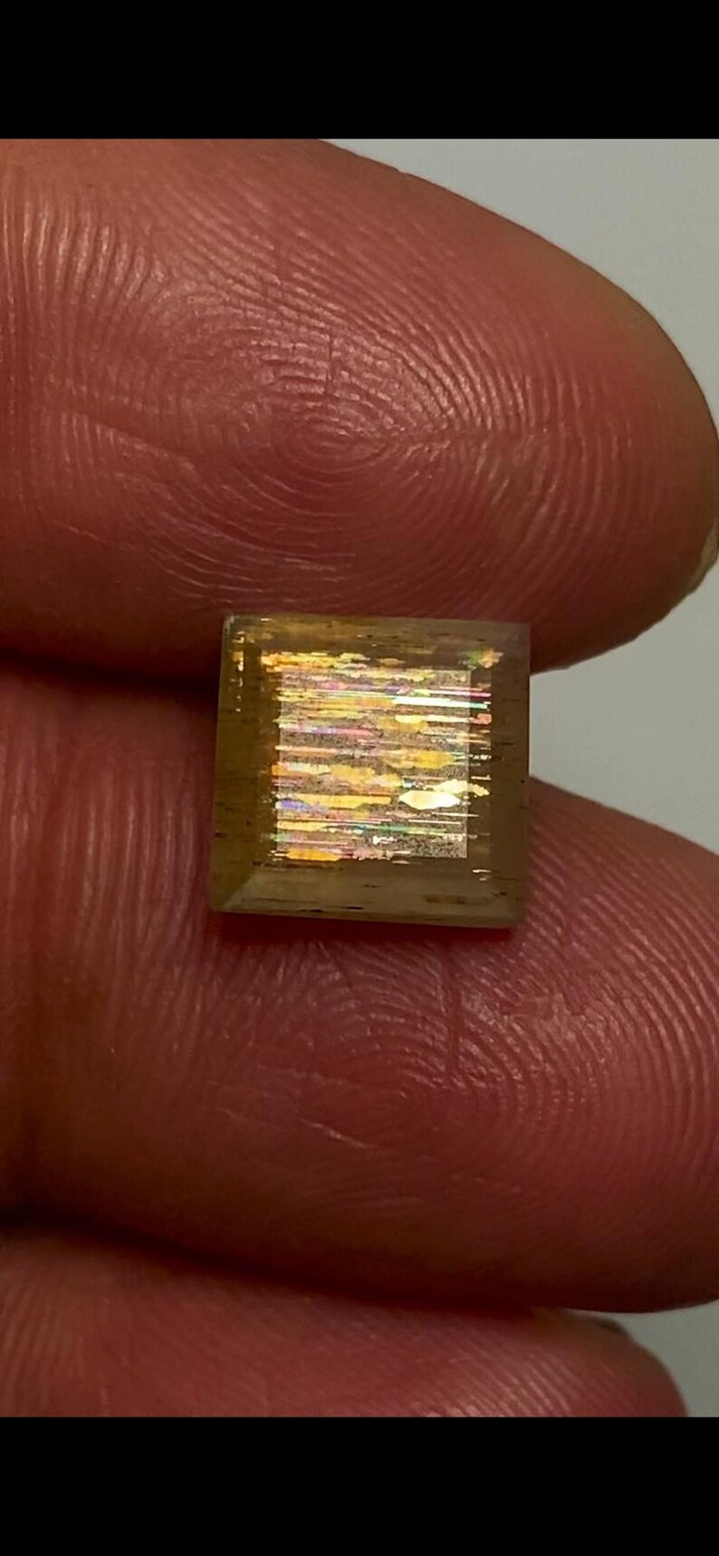 May include: A square-cut sunstone gemstone featuring a translucent brownish-orange body with internal metallic inclusions that create a shimmering, multi-coloured iridescent effect across the centre of the stone's flat, polished surface.