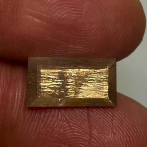 May include: A rectangular, faceted gemstone with a translucent brown body and a central, shimmering metallic inclusion that displays iridescent flashes of gold, orange, and green light across its flat, polished surface.