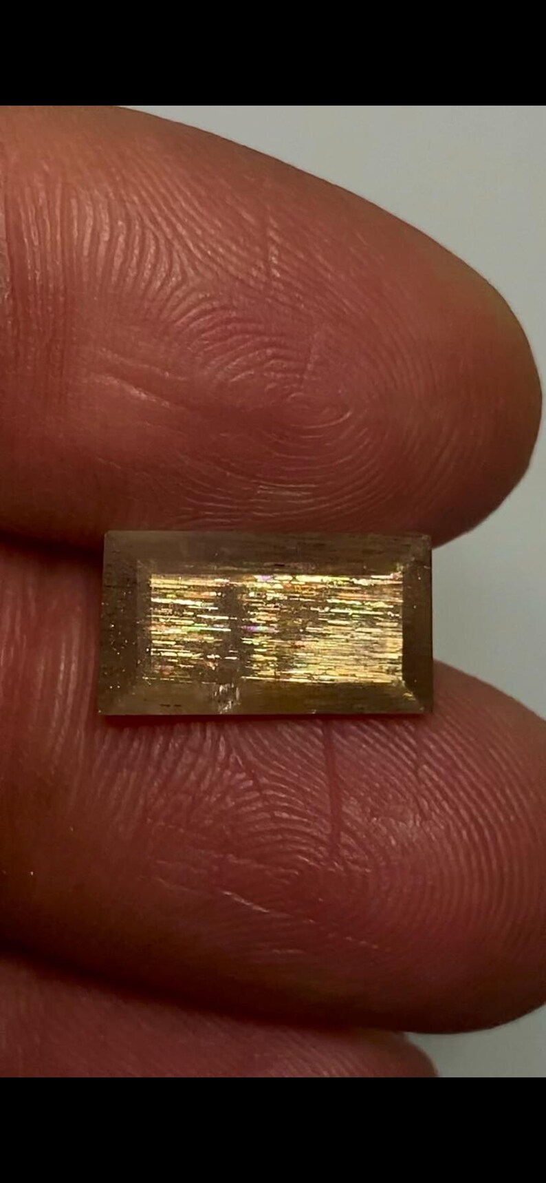 May include: A rectangular, faceted gemstone with a translucent brown body and a central, shimmering metallic inclusion that displays iridescent flashes of gold, orange, and green light across its flat, polished surface.