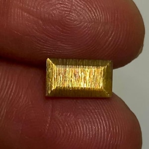May include: A rectangular, faceted sunstone gemstone with a golden-yellow hue and a distinct shimmering, metallic internal lustre. The stone features a flat top surface with beveled edges, displaying a reflective, fibrous texture throughout its body.