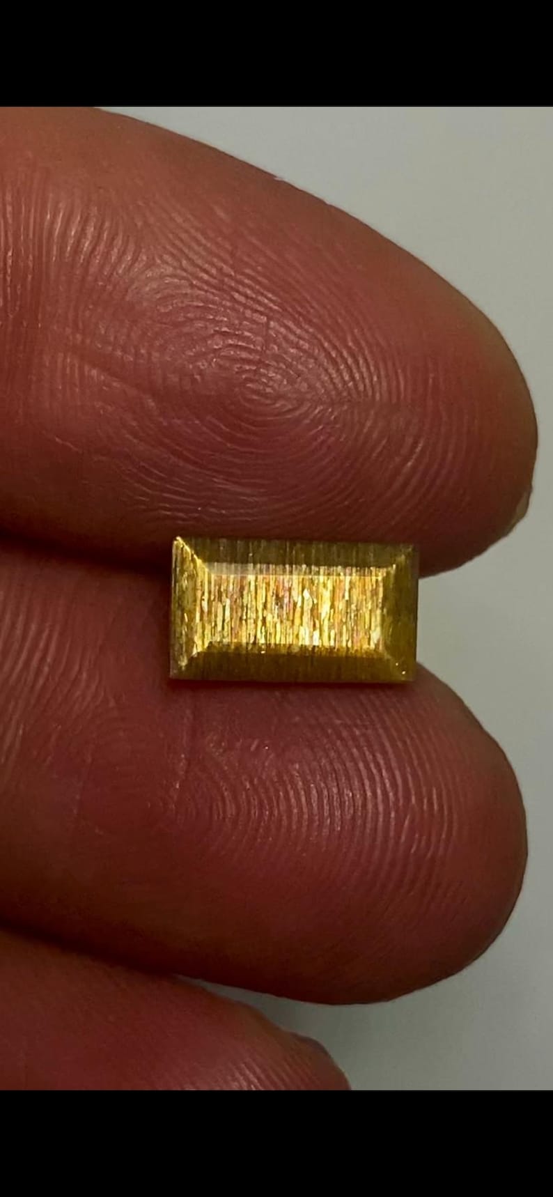 May include: A rectangular, faceted sunstone gemstone with a golden-yellow hue and a distinct shimmering, metallic internal lustre. The stone features a flat top surface with beveled edges, displaying a reflective, fibrous texture throughout its body.