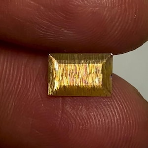 May include: A rectangular sunstone gemstone with a faceted cut, displaying a golden-yellow hue and a shimmering, metallic aventurescence effect across its surface, characterised by fine, parallel internal inclusions that catch the light.