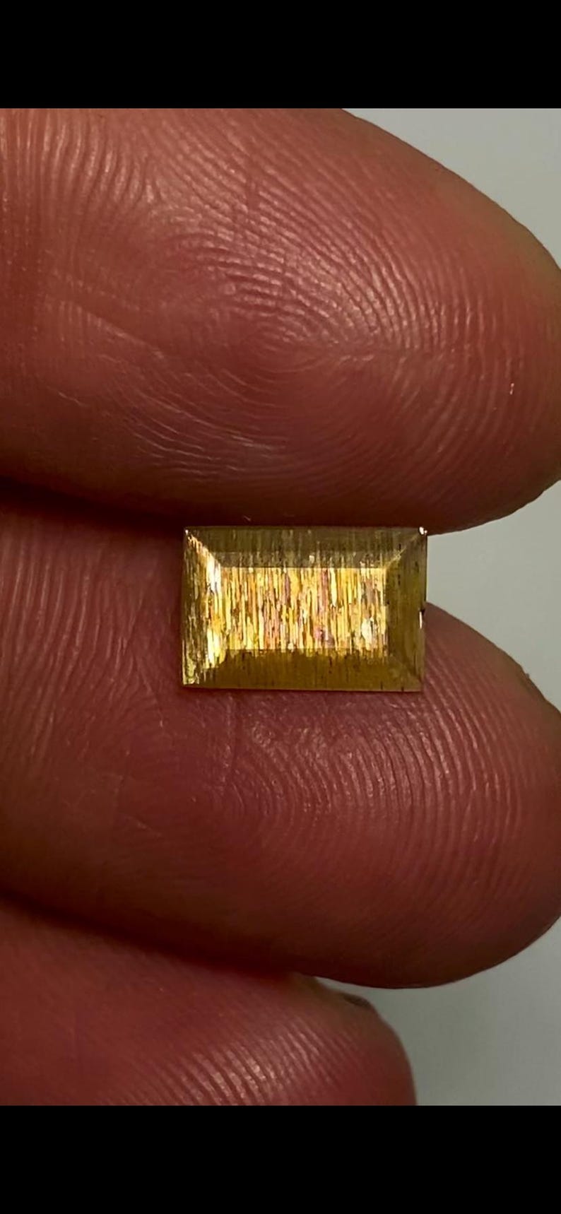 May include: A rectangular sunstone gemstone with a faceted cut, displaying a golden-yellow hue and a shimmering, metallic aventurescence effect across its surface, characterised by fine, parallel internal inclusions that catch the light.