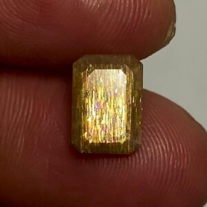 May include: A rectangular faceted sunstone gemstone with a golden-brown hue and internal metallic aventurescence displaying shimmering flashes of orange, yellow, and pink light throughout the translucent crystalline structure of the stone.