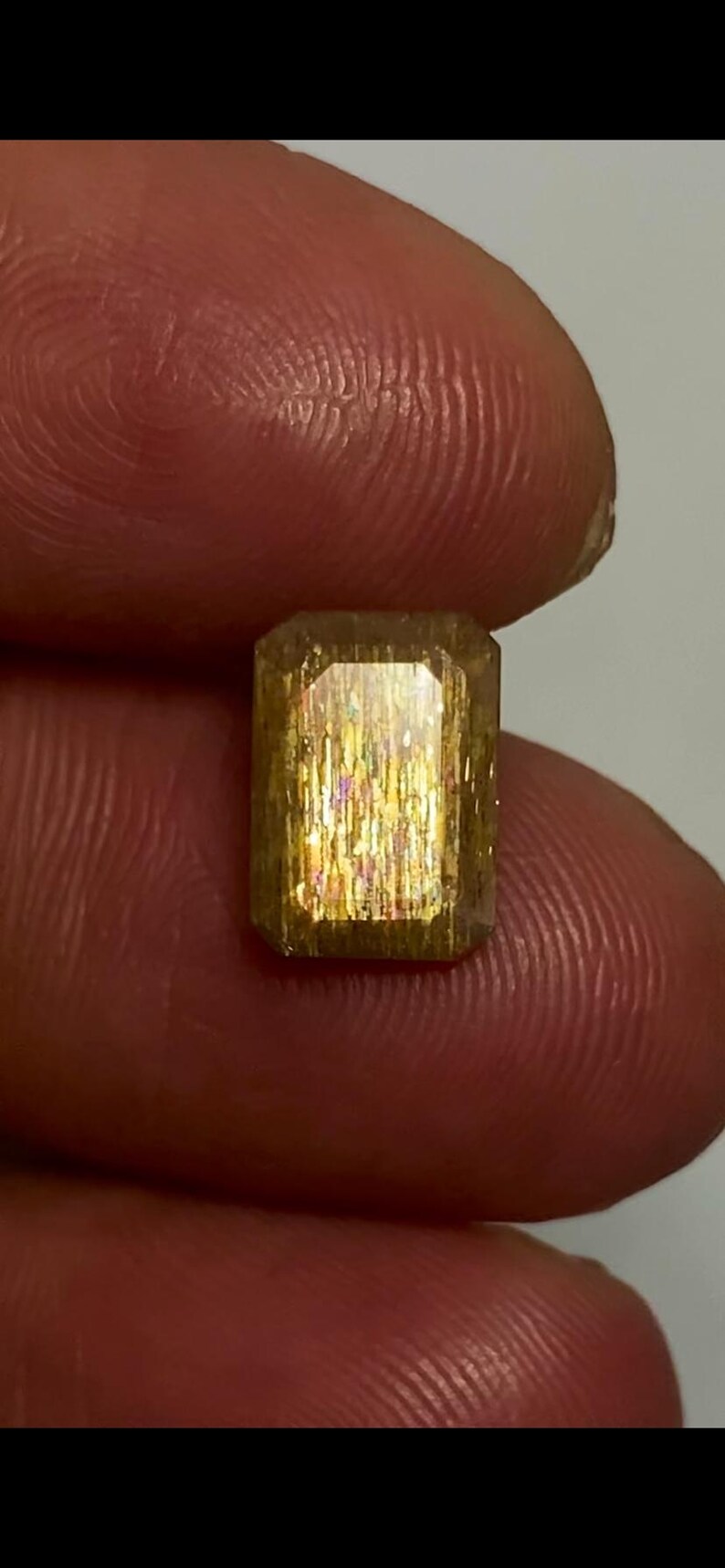 May include: A rectangular faceted sunstone gemstone with a golden-brown hue and internal metallic aventurescence displaying shimmering flashes of orange, yellow, and pink light throughout the translucent crystalline structure of the stone.