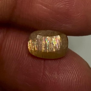 May include: A faceted oval sunstone gemstone featuring a translucent golden-brown base colour with internal metallic inclusions that create a distinct shimmering aventurescence effect with flashes of rainbow colours across the surface of the stone.