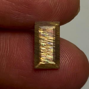 May include: A rectangular sunstone gemstone featuring a faceted surface with a golden-brown base colour and internal iridescent, metallic-like inclusions that create a shimmering, multi-coloured light reflection across the centre of the stone.