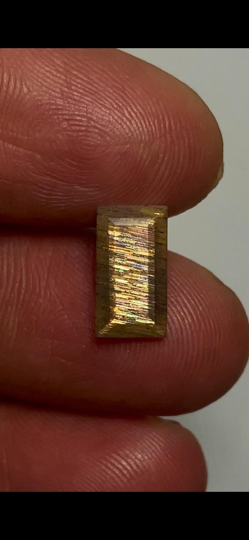 May include: A rectangular sunstone gemstone featuring a faceted surface with a golden-brown base colour and internal iridescent, metallic-like inclusions that create a shimmering, multi-coloured light reflection across the centre of the stone.