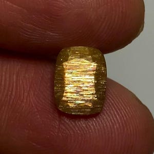 May include: A cushion-cut sunstone gemstone featuring a golden-yellow hue with internal metallic schiller effects. The stone displays a faceted surface with horizontal light reflections and a warm, shimmering appearance throughout its structure.