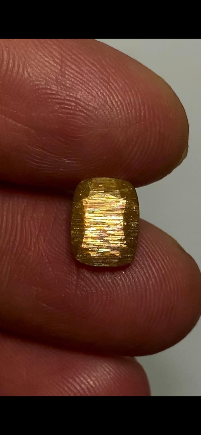 May include: A cushion-cut sunstone gemstone featuring a golden-yellow hue with internal metallic schiller effects. The stone displays a faceted surface with horizontal light reflections and a warm, shimmering appearance throughout its structure.
