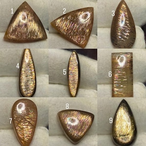 Puede incluir: Una colección de nueve piedras preciosas de piedra de sol pulidas en varias formas, incluyendo triángulos, gotas, óvalos y rectángulos. Cada piedra muestra un tono marrón dorado con aventurescencia metálica interna y reflejos iridiscentes.