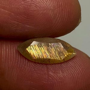 May include: A marquise-cut sunstone gemstone featuring a translucent golden-brown hue with internal metallic aventurescence, displaying shimmering streaks of orange, yellow, and green light across its faceted surface.