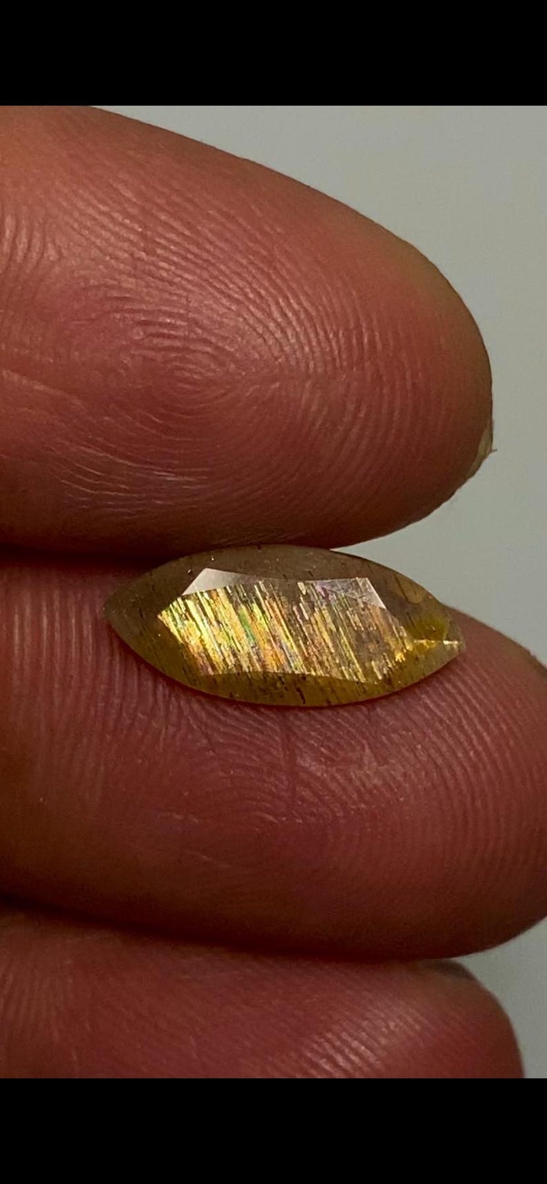 May include: A marquise-cut sunstone gemstone featuring a translucent golden-brown hue with internal metallic aventurescence, displaying shimmering streaks of orange, yellow, and green light across its faceted surface.