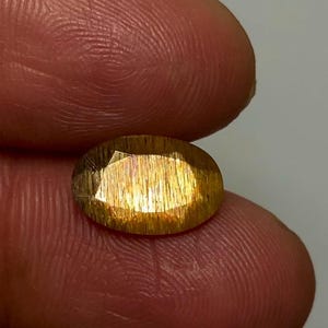 May include: An oval-cut sunstone gemstone featuring a golden-orange hue with distinct internal metallic inclusions that create a shimmering aventurescence effect across the faceted surface of the translucent, polished stone.