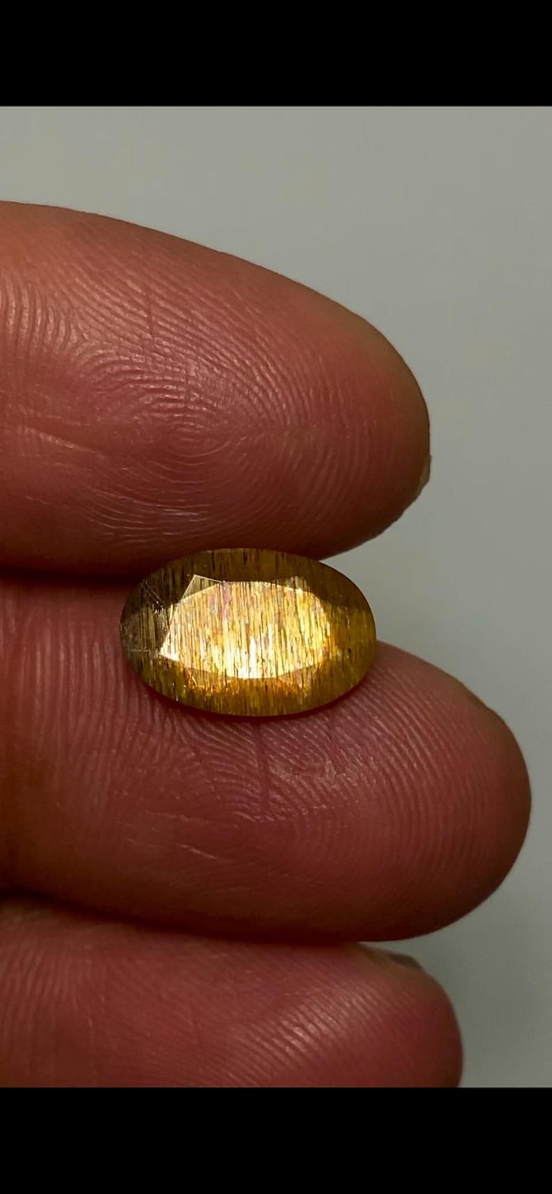 May include: An oval-cut sunstone gemstone featuring a golden-orange hue with distinct internal metallic inclusions that create a shimmering aventurescence effect across the faceted surface of the translucent, polished stone.