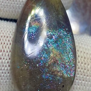 Puede incluir: Una gema pulida en forma de lágrima. La base oscura de la piedra está salpicada de inclusiones iridiscentes de arcoíris. La parte superior de la piedra tiene un tono rojizo.