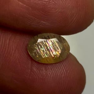 May include: An oval-shaped sunstone gemstone featuring a translucent golden-brown body with distinct internal metallic inclusions that reflect iridescent rainbow colours, including flashes of pink, green, and yellow across its faceted surface.