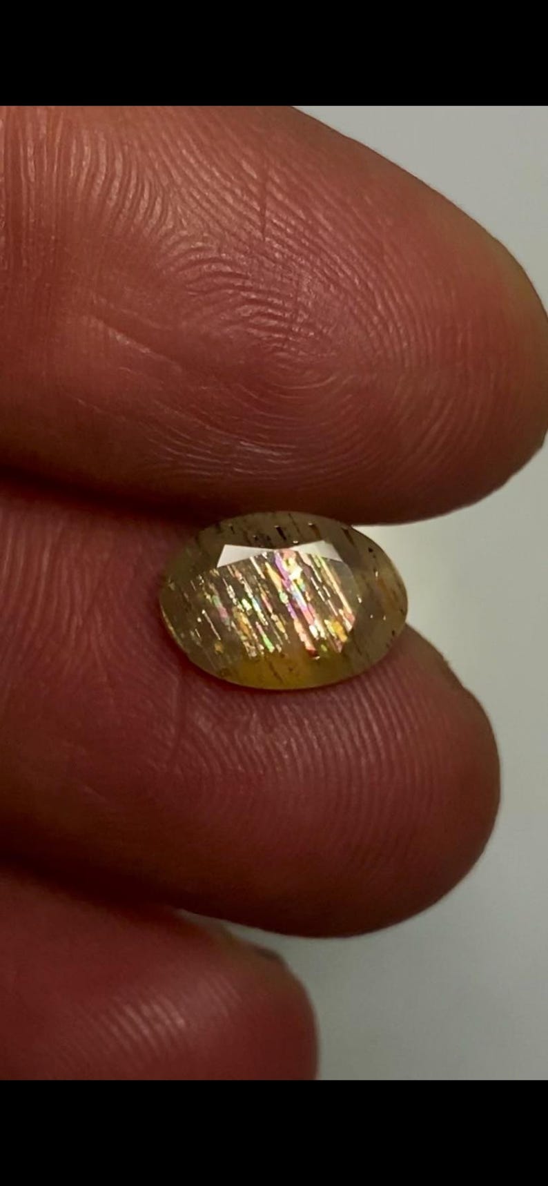 May include: An oval-shaped sunstone gemstone featuring a translucent golden-brown body with distinct internal metallic inclusions that reflect iridescent rainbow colours, including flashes of pink, green, and yellow across its faceted surface.