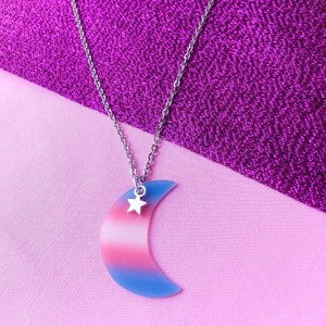 May include: A silver chain necklace featuring a crescent moon pendant. The moon is striped with pink, white, and blue, with a small silver star charm. The necklace is set against a pink and purple backdrop, creating a striking contrast.